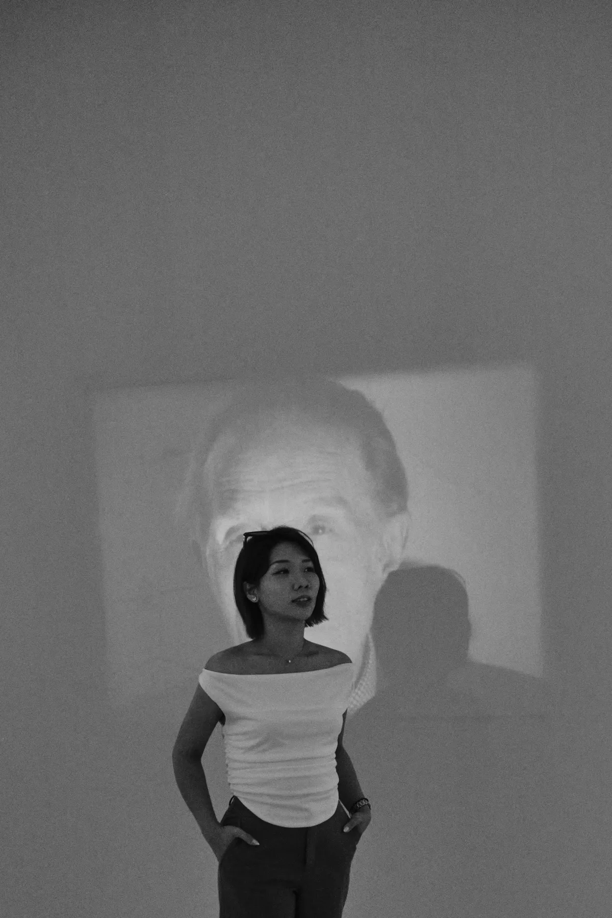 Black-and-white photo of Janus, hands in pockets, standing in front of a large projected portrait
